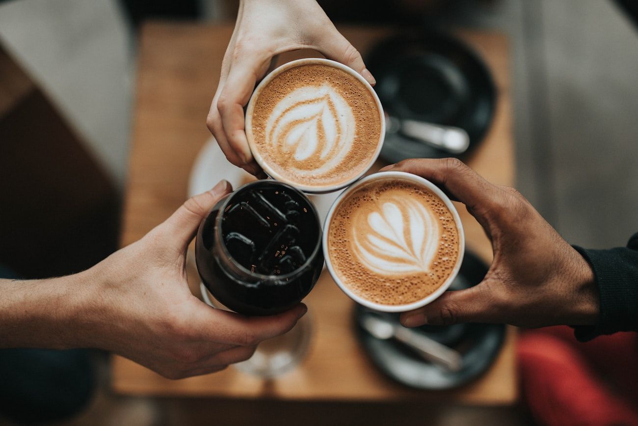 Three hands raising lattes with latte art over a wooden café table.