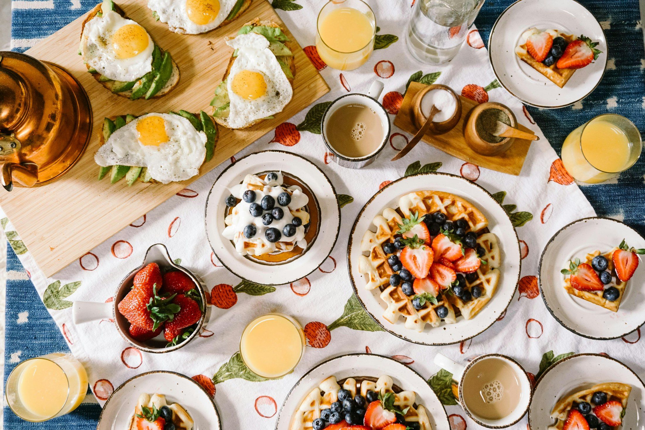 An overhead spread of breakfast plates — waffles, eggs, fresh berries, coffee and juice — on a bright table.